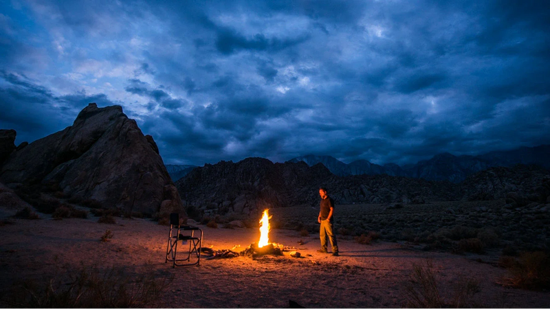 Woman holding camp cup by fire with mountains in background.