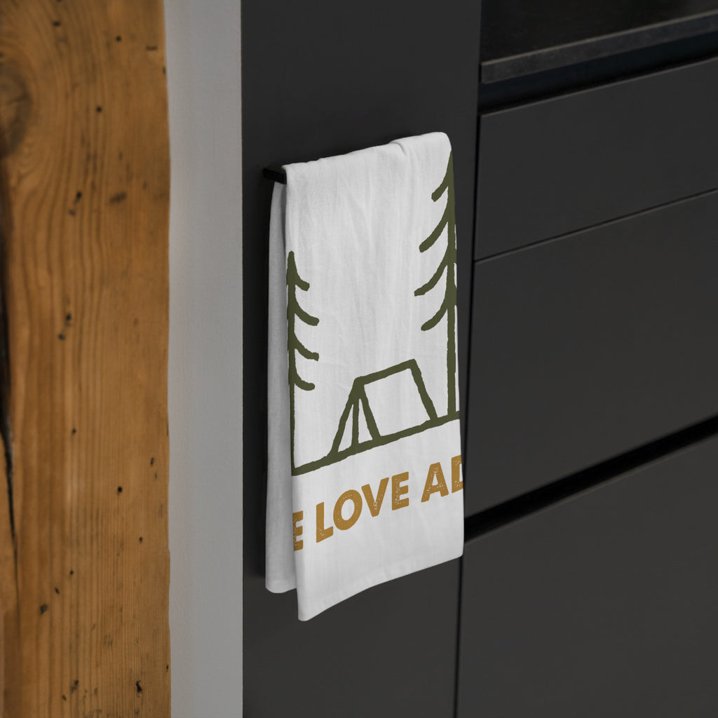 White towel with green and gold WE LOVE ADV camp design hanging on a dark grey surface.